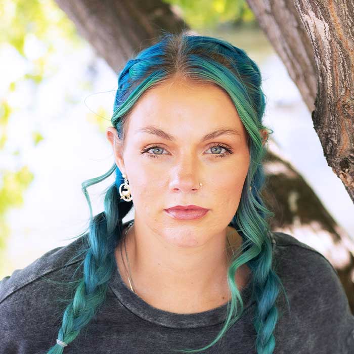 Woman with teal-colored wavy hair wearing black shirt, earrings, outdoors under tree