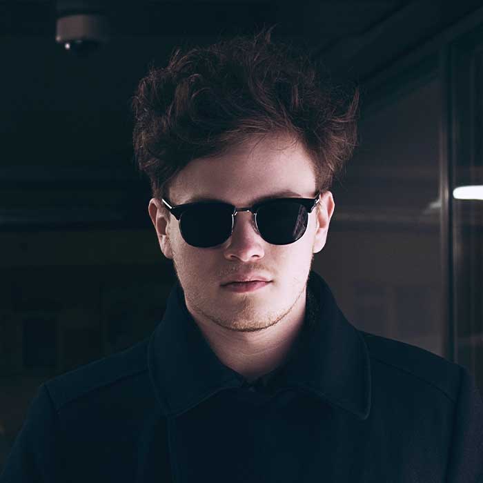 Man wearing dark sunglasses and black jacket in dimly lit indoor setting, looking at camera